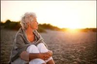 A woman sitting on the beach looking out at the sunset.